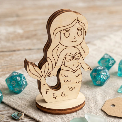 Detailed close-up of a laser cut plywood mermaid figurine with wood grain layers, standing on linen near teal glittery dice.