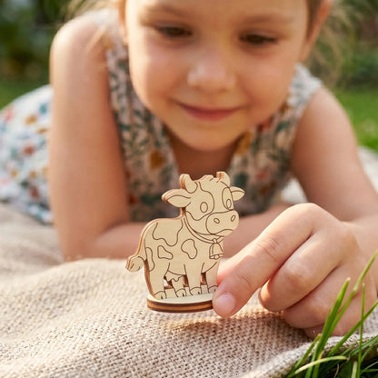 Child looking at a tiny cow figurine made of laser cut plywood, showing its scale on a rustic blanket.
