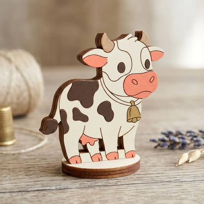 A hand-painted spotted miniature cow figurine with a bell, made of laser cut plywood and staged on a wooden table.
