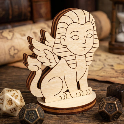 Close-up of a laser cut plywood sphinx figurine on a fantasy map with beetle-etched dice and a vintage hourglass backdrop.
