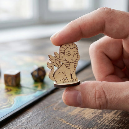 Adult hand holding a tiny laser cut plywood sphinx figurine to show scale, with a tabletop RPG map in the blurry background