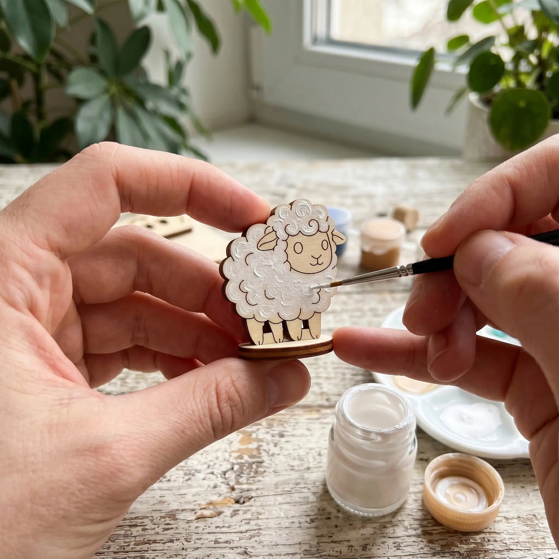 Close-up of hands using a fine brush to paint white acrylic onto a miniature laser cut plywood sheep toy on a work table.