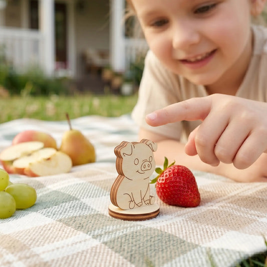 Miniature laser cut plywood pig figurine on a picnic blanket next to fruit, with a smiling child pointing at it.