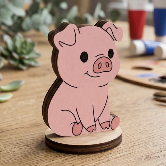 A hand-painted pink miniature pig figurine made of laser cut plywood, staged on a wooden art desk with acrylic paint tubes