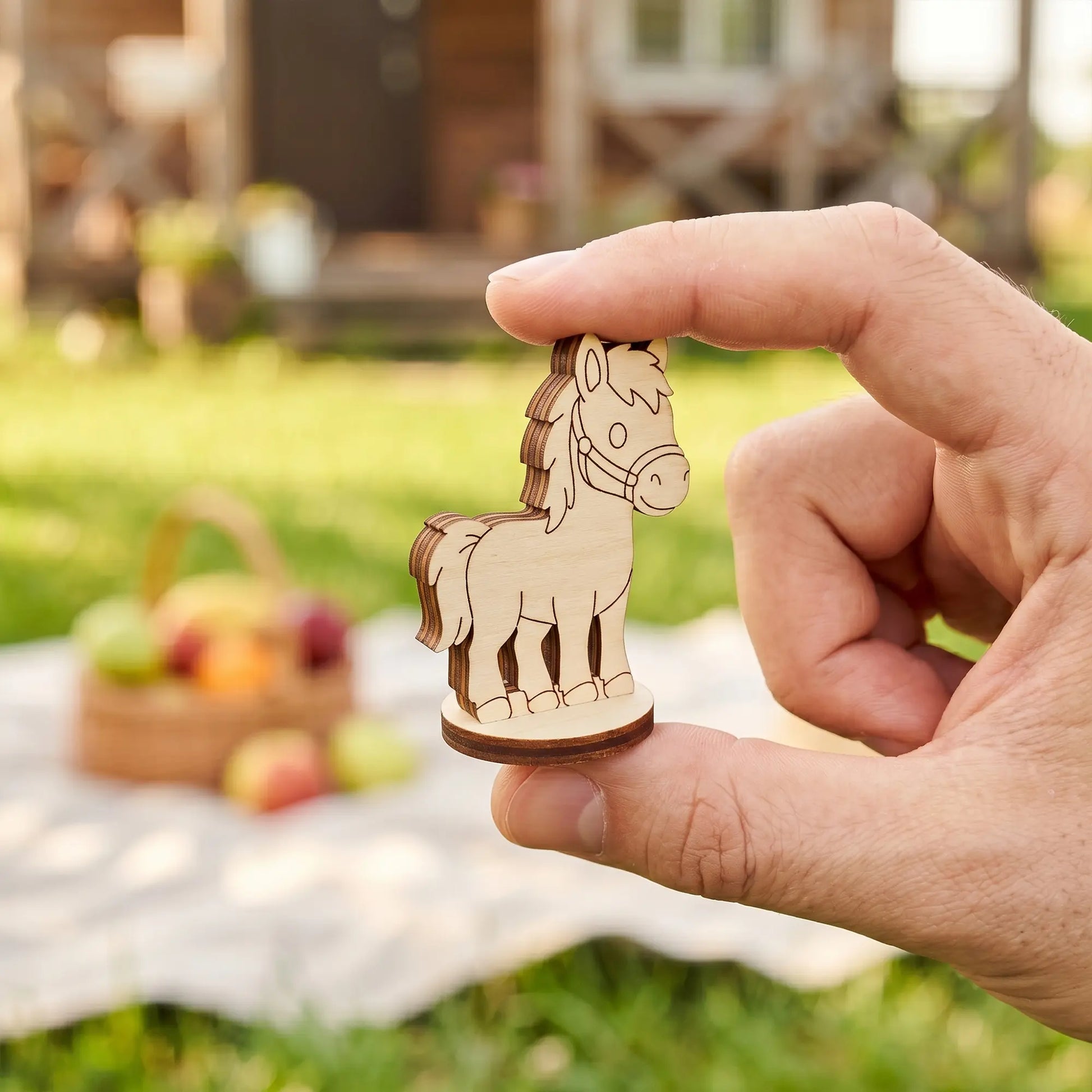 Close-up showing fingers holding a miniature standing horse figurine made of laser cut plywood over a picnic setup.
