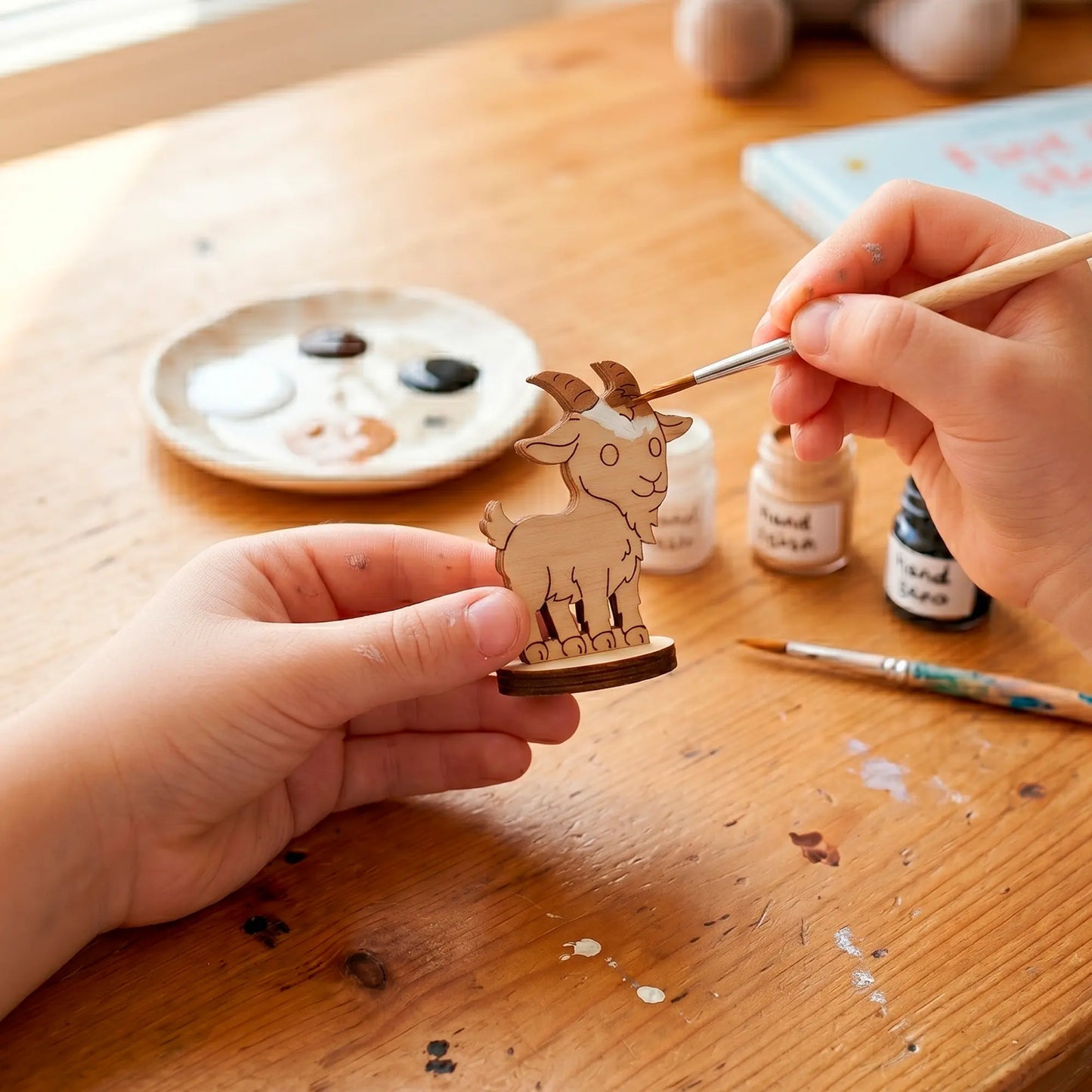 Close-up of hands using a fine brush to paint white acrylic onto a miniature laser cut plywood goat toy at a desk.