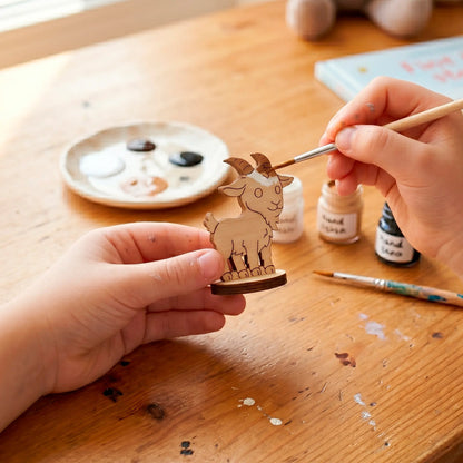 Close-up of hands using a fine brush to paint white acrylic onto a miniature laser cut plywood goat toy at a desk.