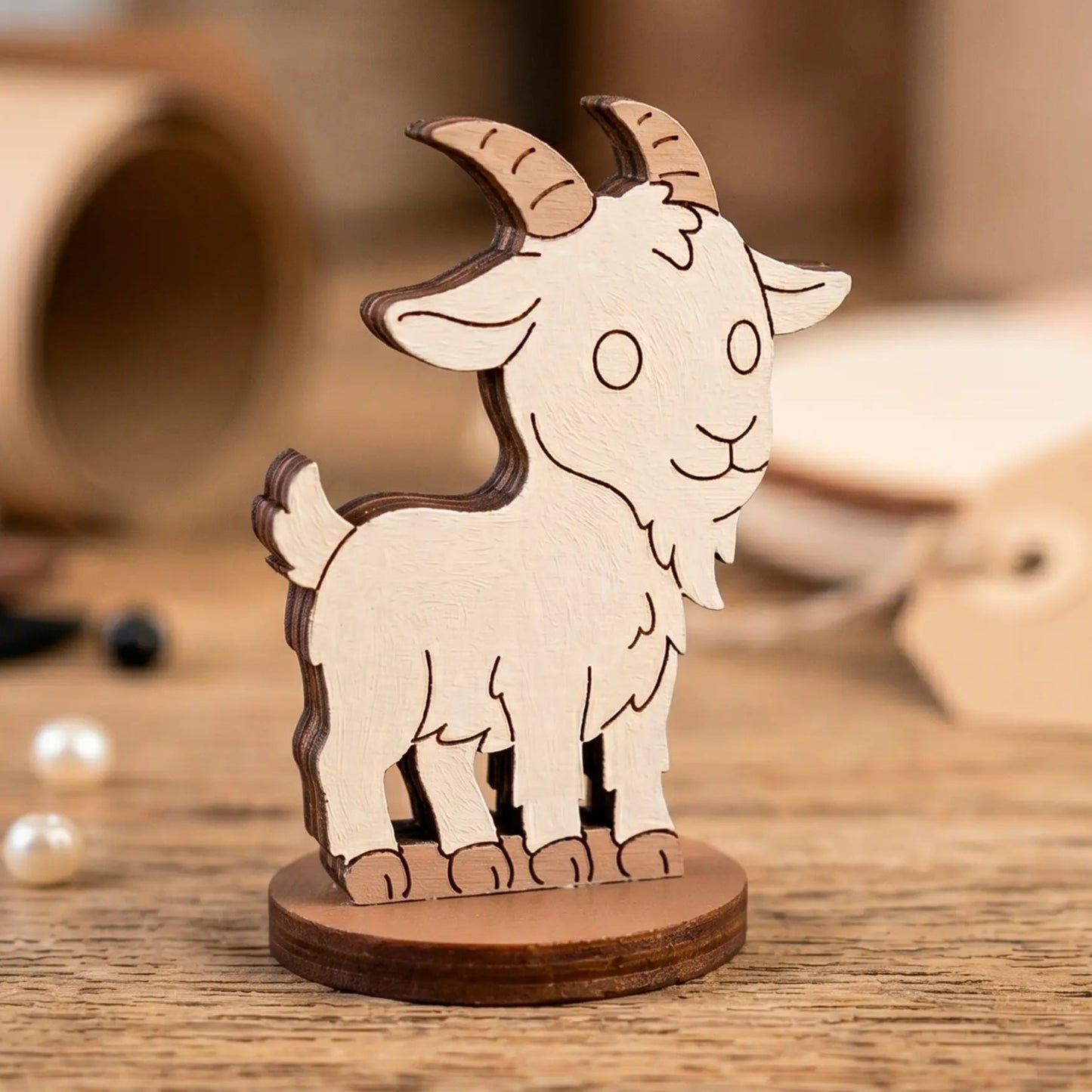 A hand-painted white miniature goat figurine with horns, made of laser cut plywood and staged on a wooden workshop desk.