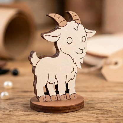 A hand-painted white miniature goat figurine with horns, made of laser cut plywood and staged on a wooden workshop desk.