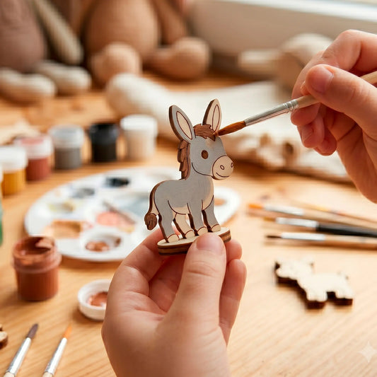 Close-up of hands using a fine brush to paint grey acrylic onto a miniature laser cut plywood donkey toy at a desk.
