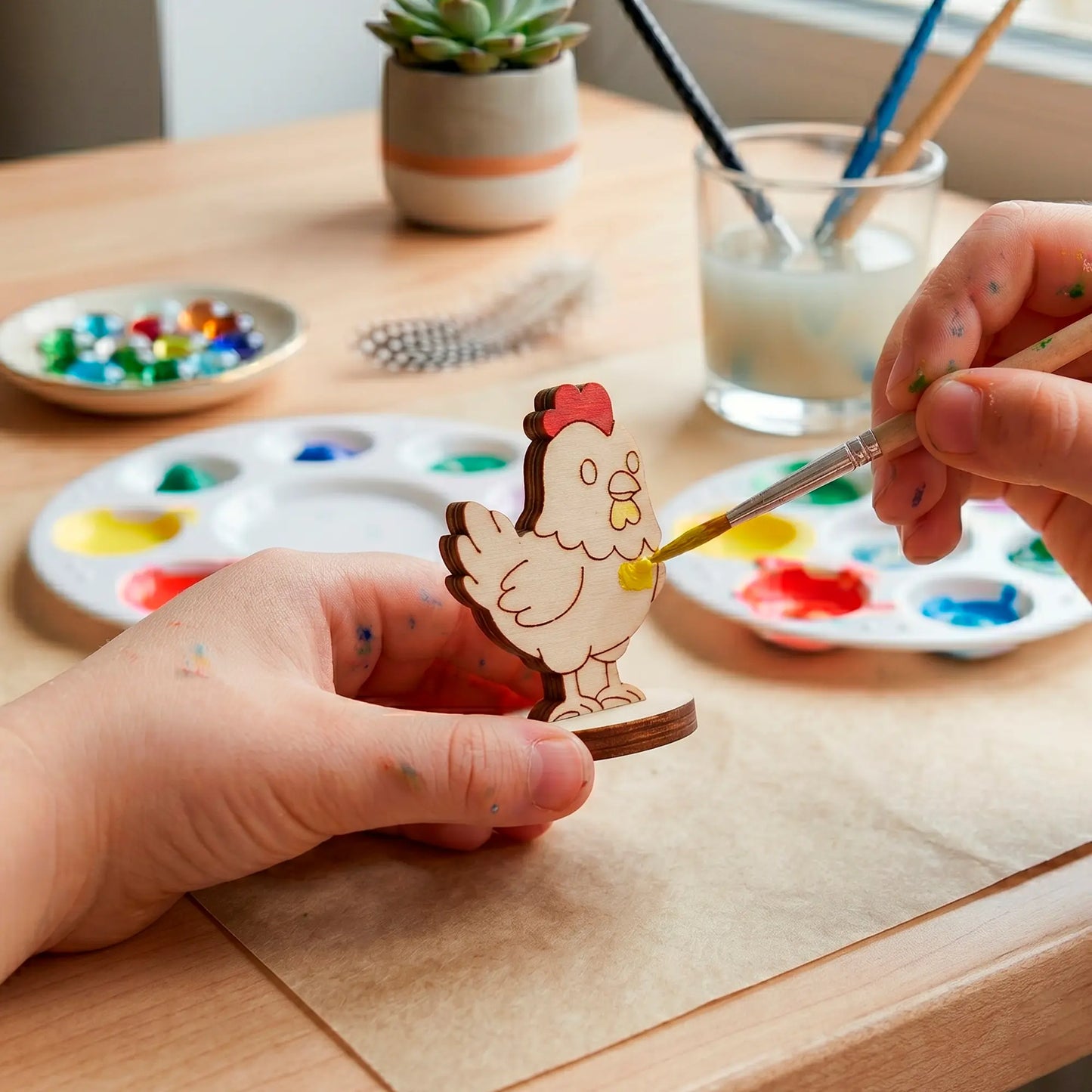 Child hands painting a laser cut plywood cute hen figure with a brush and yellow acrylic color at a wooden craft table