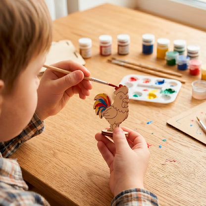 Child hands coloring a small laser cut plywood cute rooster figurine with red acrylic paint at a wooden craft desk.