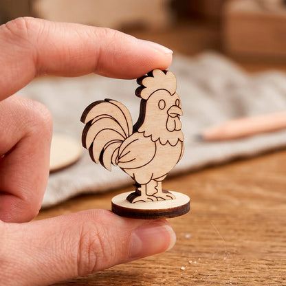 Two fingers holding a tiny laser cut plywood cute rooster figure, showing its miniature scale over a blurry workshop
