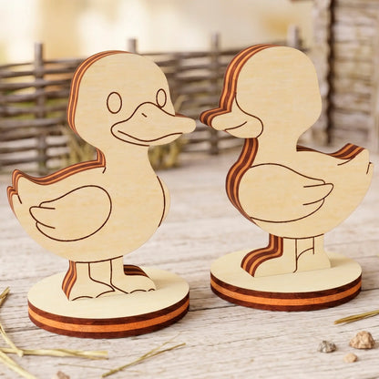 Two laser cut plywood cute duck figures with neat layered edges standing on wood near a miniature wicker fence.