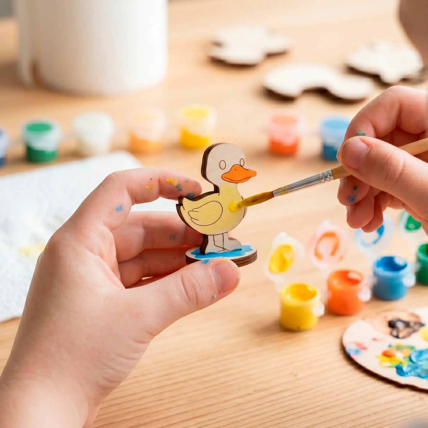 Child hands coloring a small laser cut plywood cute duck figure with yellow acrylic paint using a brush at a craft desk
