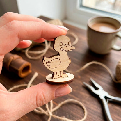 Two fingers holding a tiny laser cut plywood cute duck miniature, showing small scale over a dark wooden workshop table