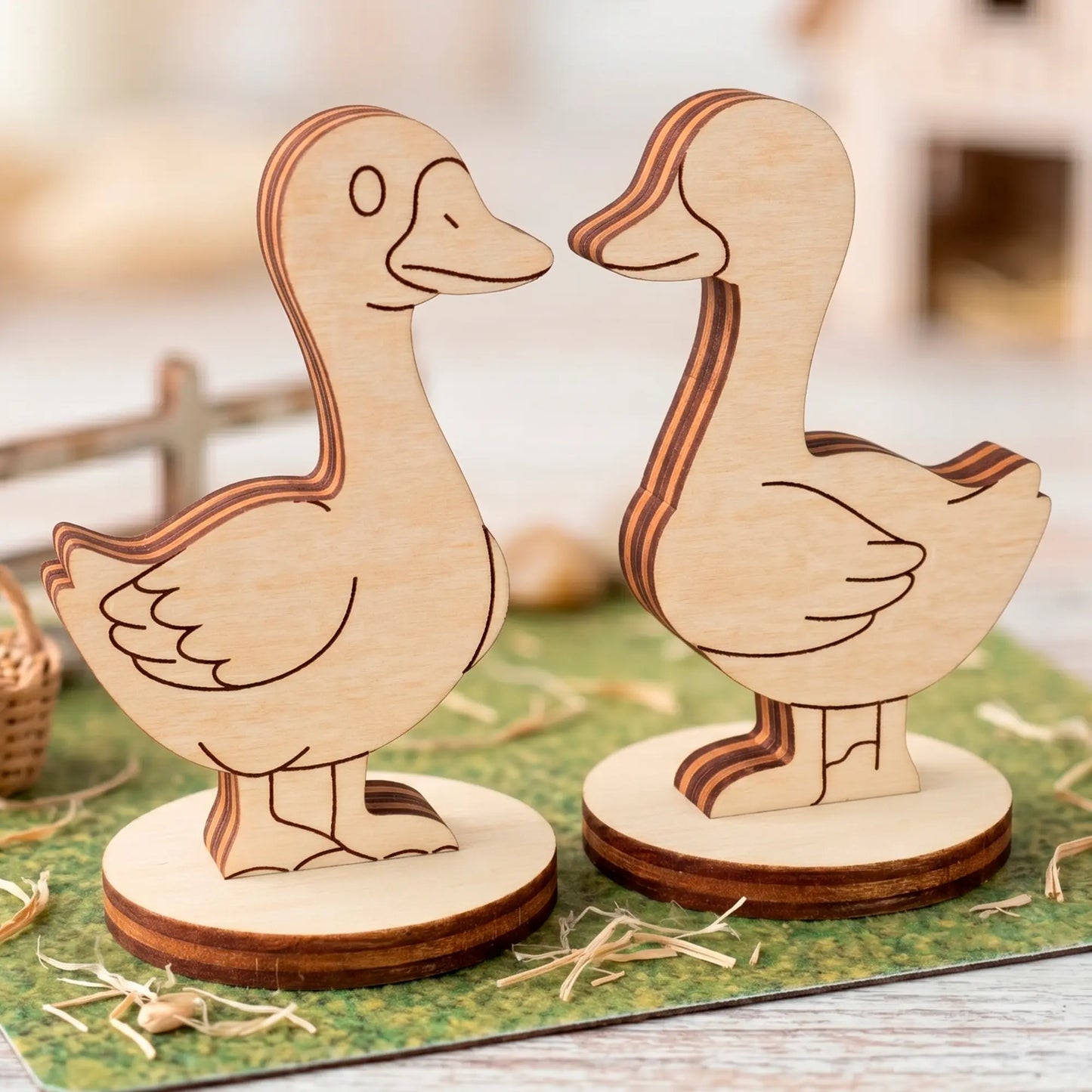 Two laser cut plywood goose figurines standing face to face on round bases over a green surface with decorative straw.