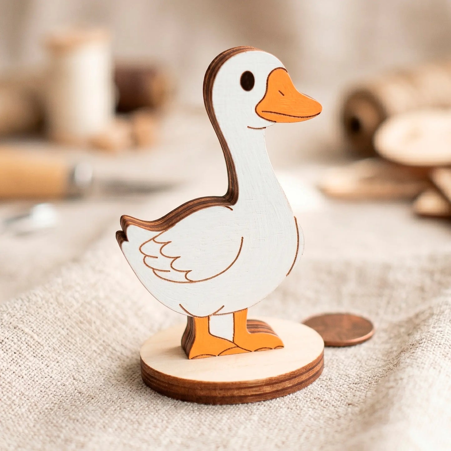 Painted laser cut plywood goose toy with a white body and orange beak standing on a linen cloth.