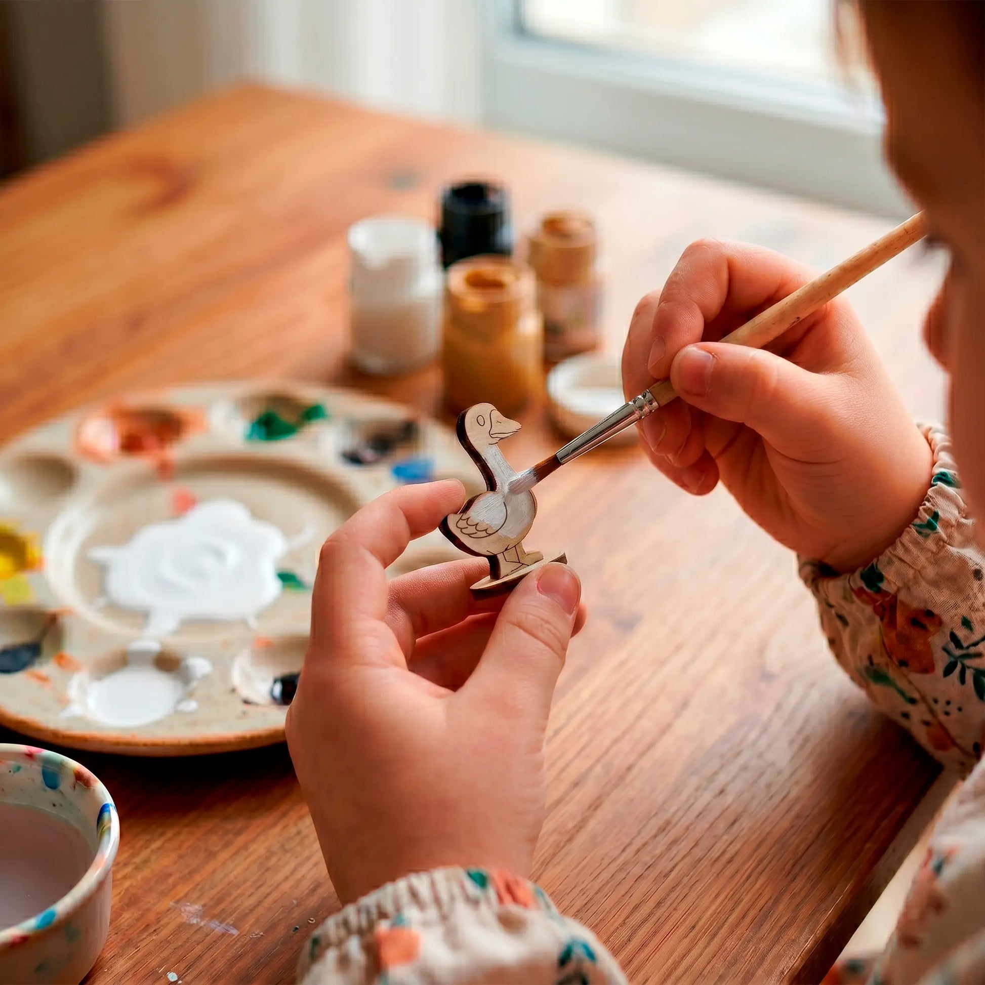 Close-up of a child painting a small laser cut plywood goose figurine with white paint using a brush at a wooden table.