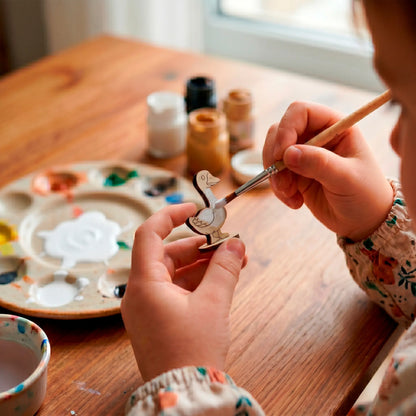 Close-up of a child painting a small laser cut plywood goose figurine with white paint using a brush at a wooden table.