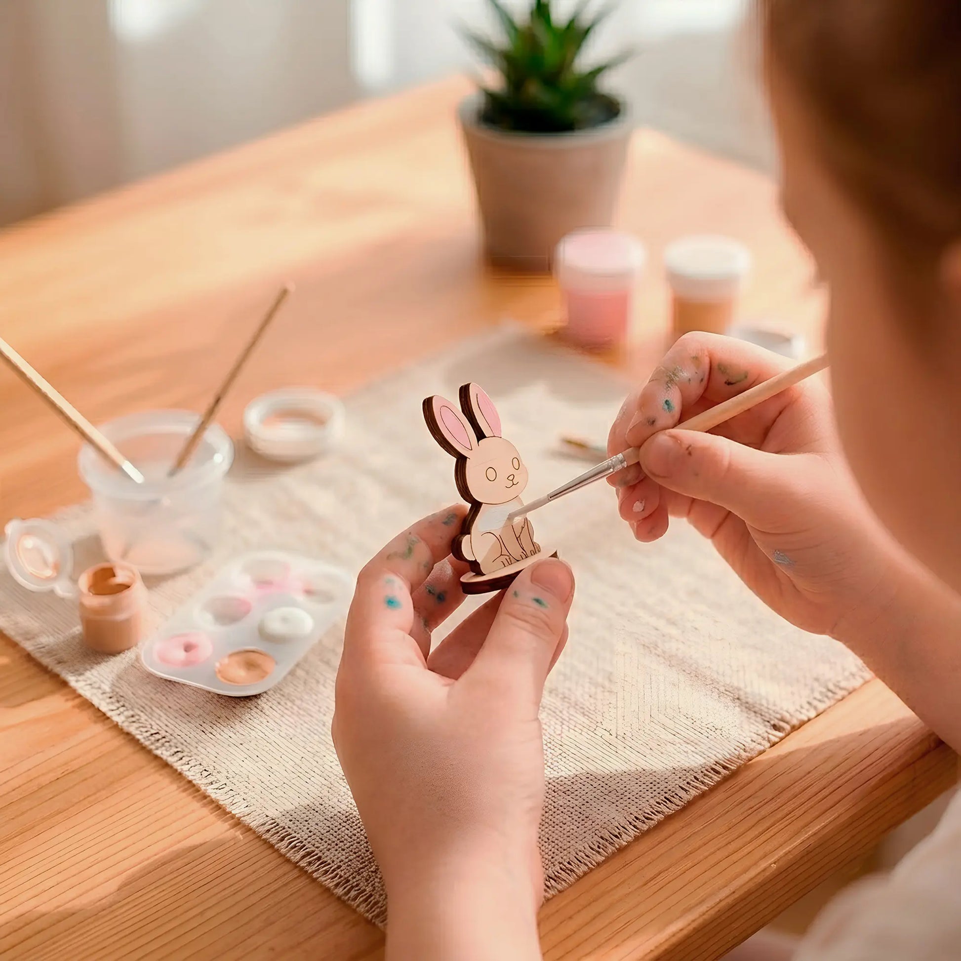 Close-up of hands painting a small laser cut plywood rabbit figurine with a fine brush at a table with paint jars.