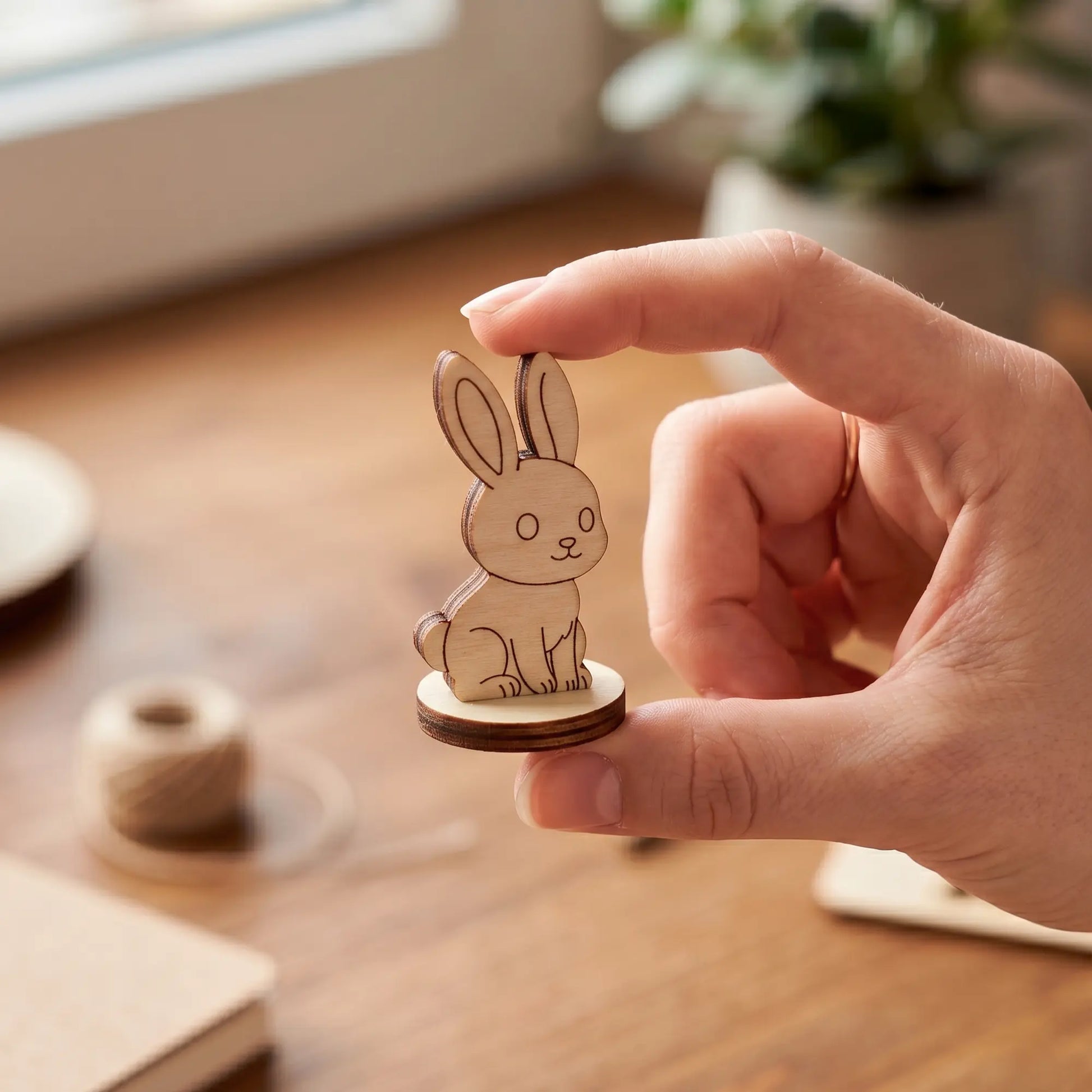 A person gently holding a miniature laser cut plywood rabbit figurine between their fingers over a cozy wooden desk.