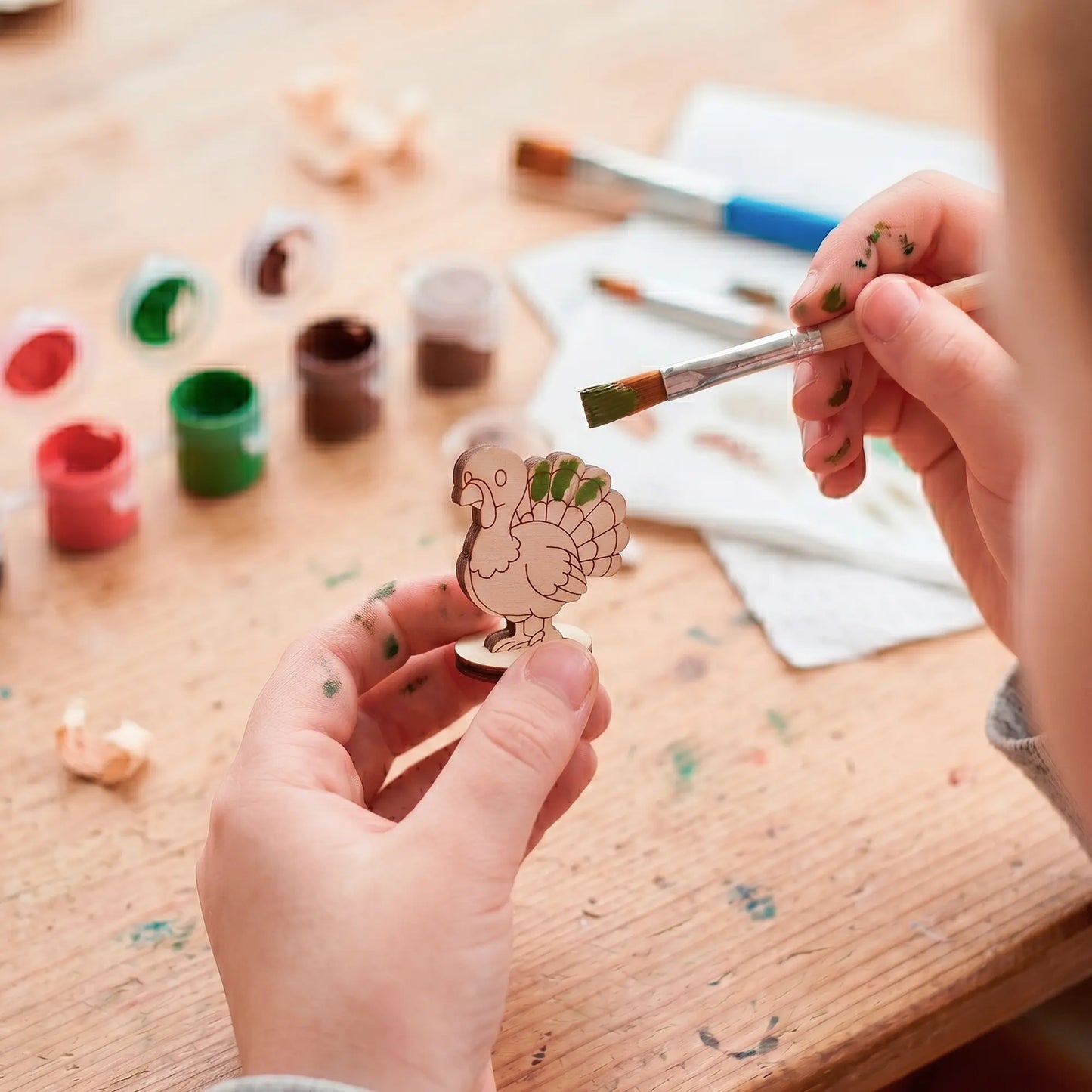 Overhead view of hands painting the feathers of a small laser cut plywood turkey toy with a brush and acrylic paints.