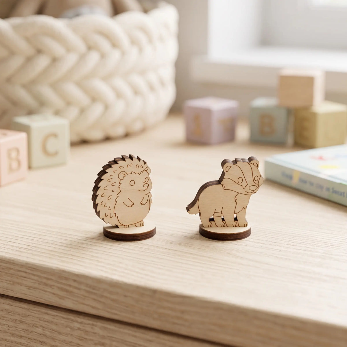Close-up of laser cut plywood hedgehog and badger figurines on round bases standing beside pastel wooden toy blocks.