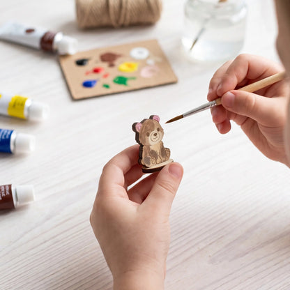 Hands paint details on laser cut plywood bear cub figurine with fine brush using acrylic paints on white table.