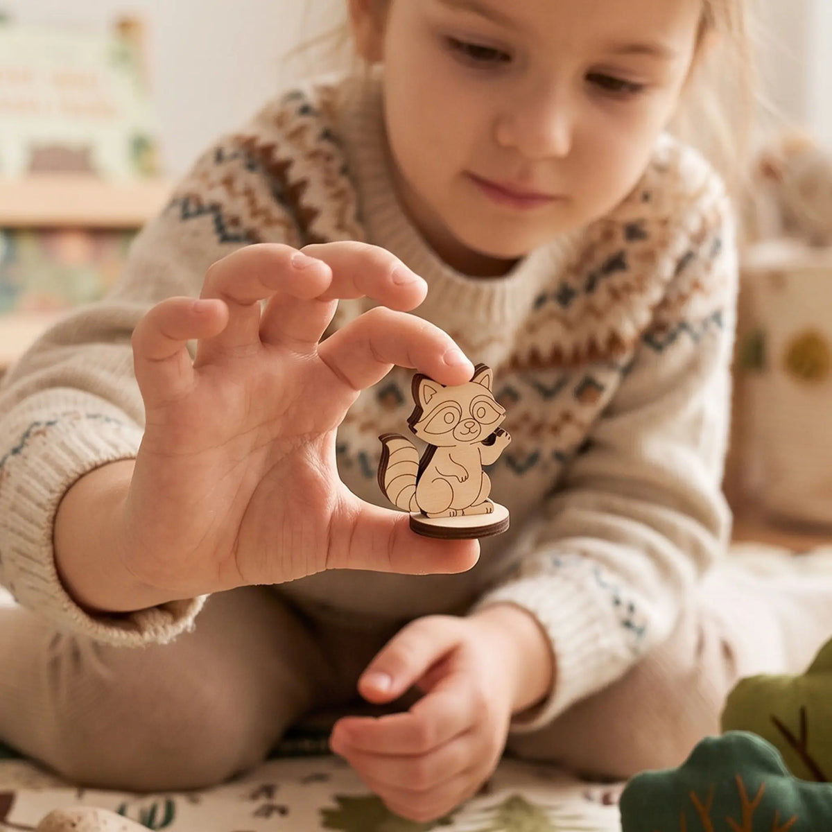 Child holds laser cut plywood raccoon figurine showing scale in cozy nursery play area with felt woodland toys.