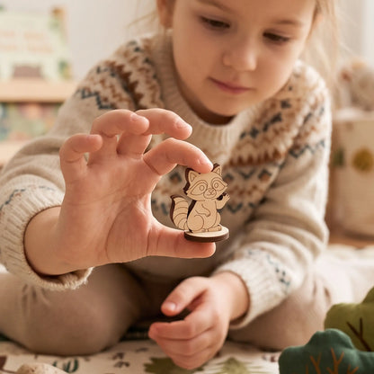 Child holds laser cut plywood raccoon figurine showing scale in cozy nursery play area with felt woodland toys.