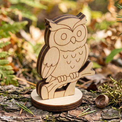 Laser cut plywood owl figurine perched on branch stand displayed outdoors on tree bark with moss and pine needles.