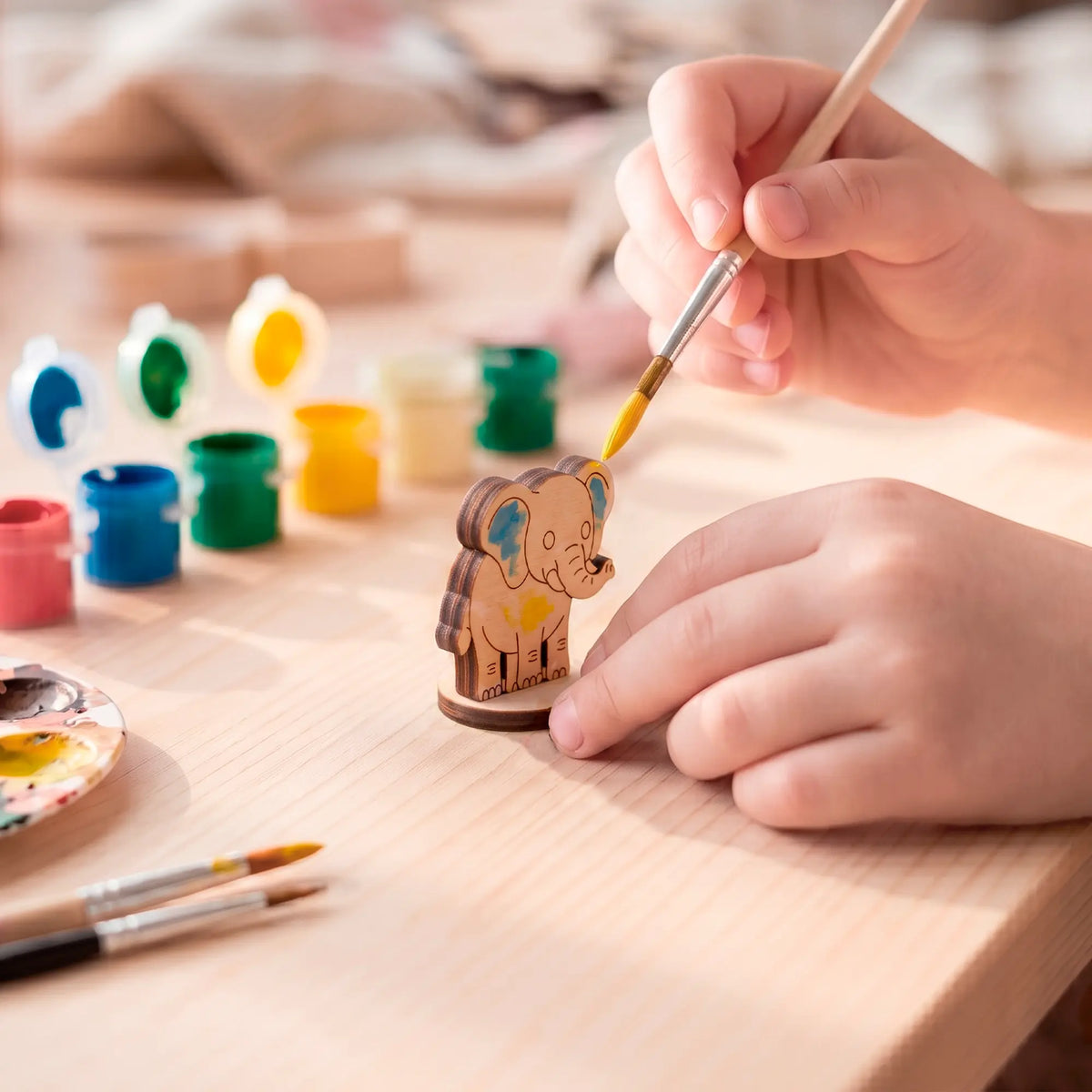 A hand using a fine paintbrush to paint a detailed laser-cut plywood elephant figurine on a wooden table with paint pots
