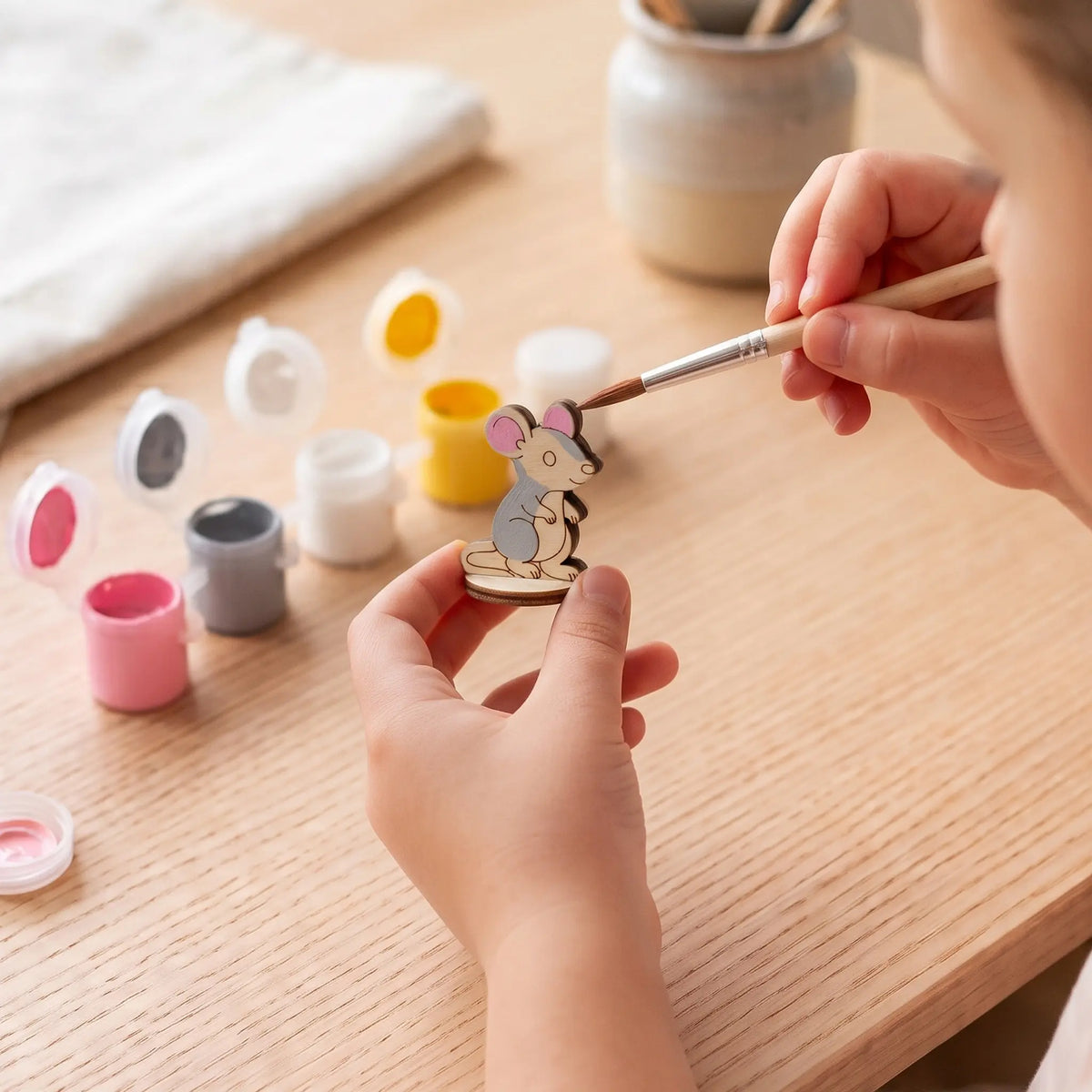 Child hand paints miniature laser cut plywood mouse figurine with fine brush using small acrylic paint pots on desk.
