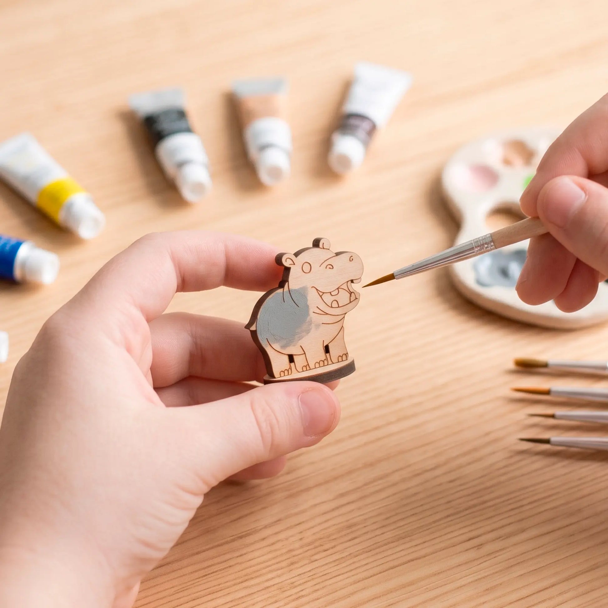 A pair of human hands painting a small laser-cut plywood hippo figurine with a fine brush surrounded by multiple paint tubes.