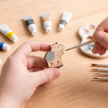 A pair of human hands painting a small laser-cut plywood hippo figurine with a fine brush surrounded by multiple paint tubes.
