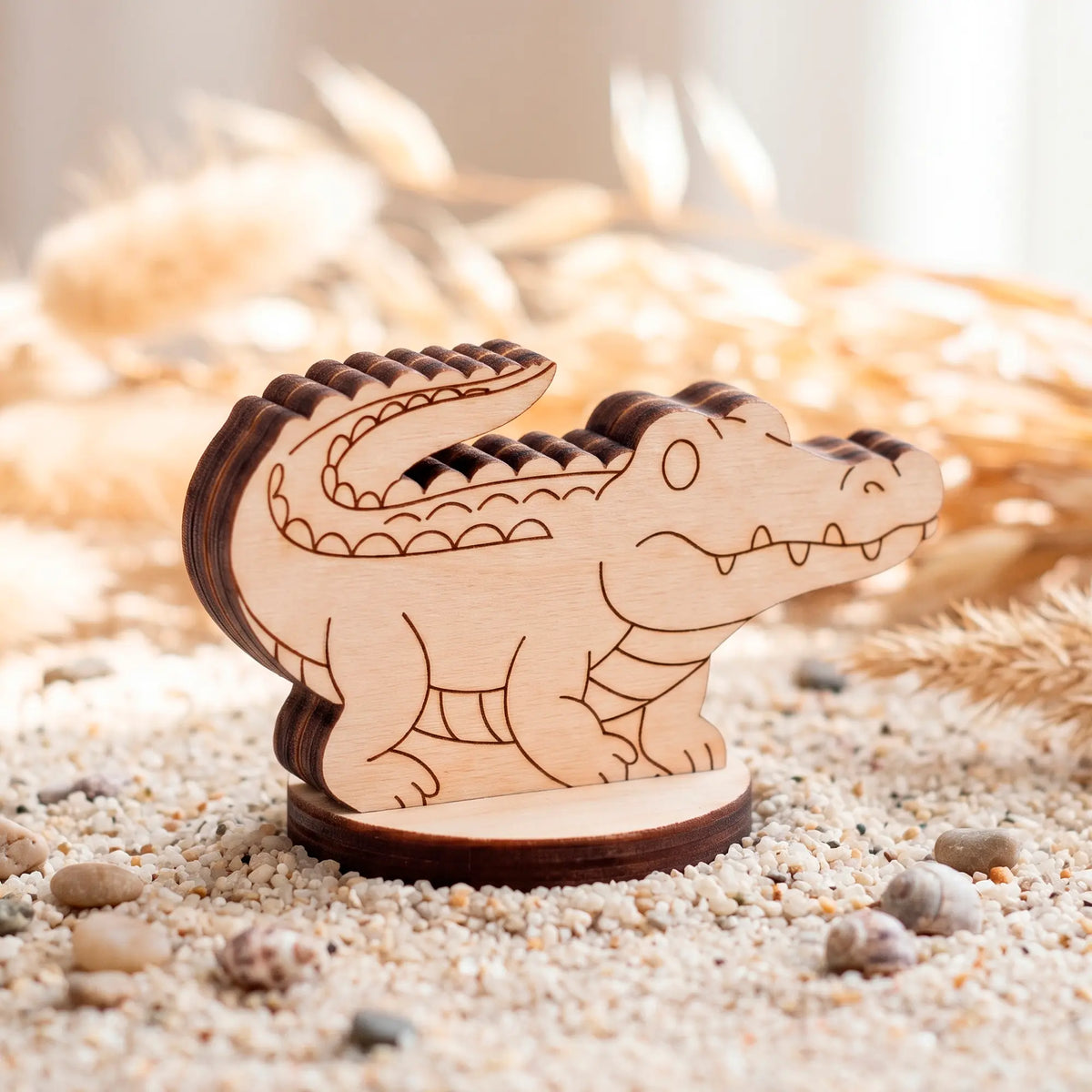 A small detailed laser-cut plywood crocodile figurine resting on a round wooden base, staged beautifully on sandy ground.