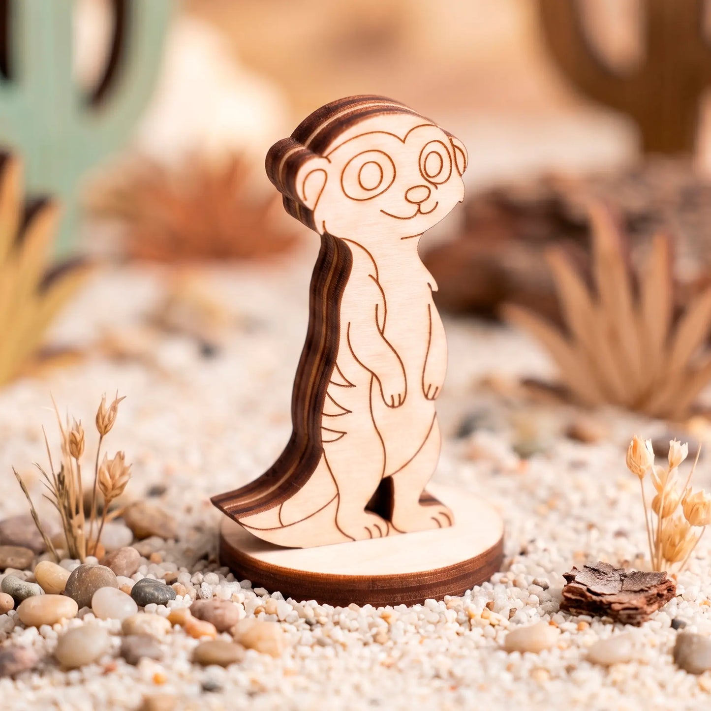 Laser-cut plywood meerkat figurine standing on a round wooden base, staged on sandy ground with pebbles and dry grass.