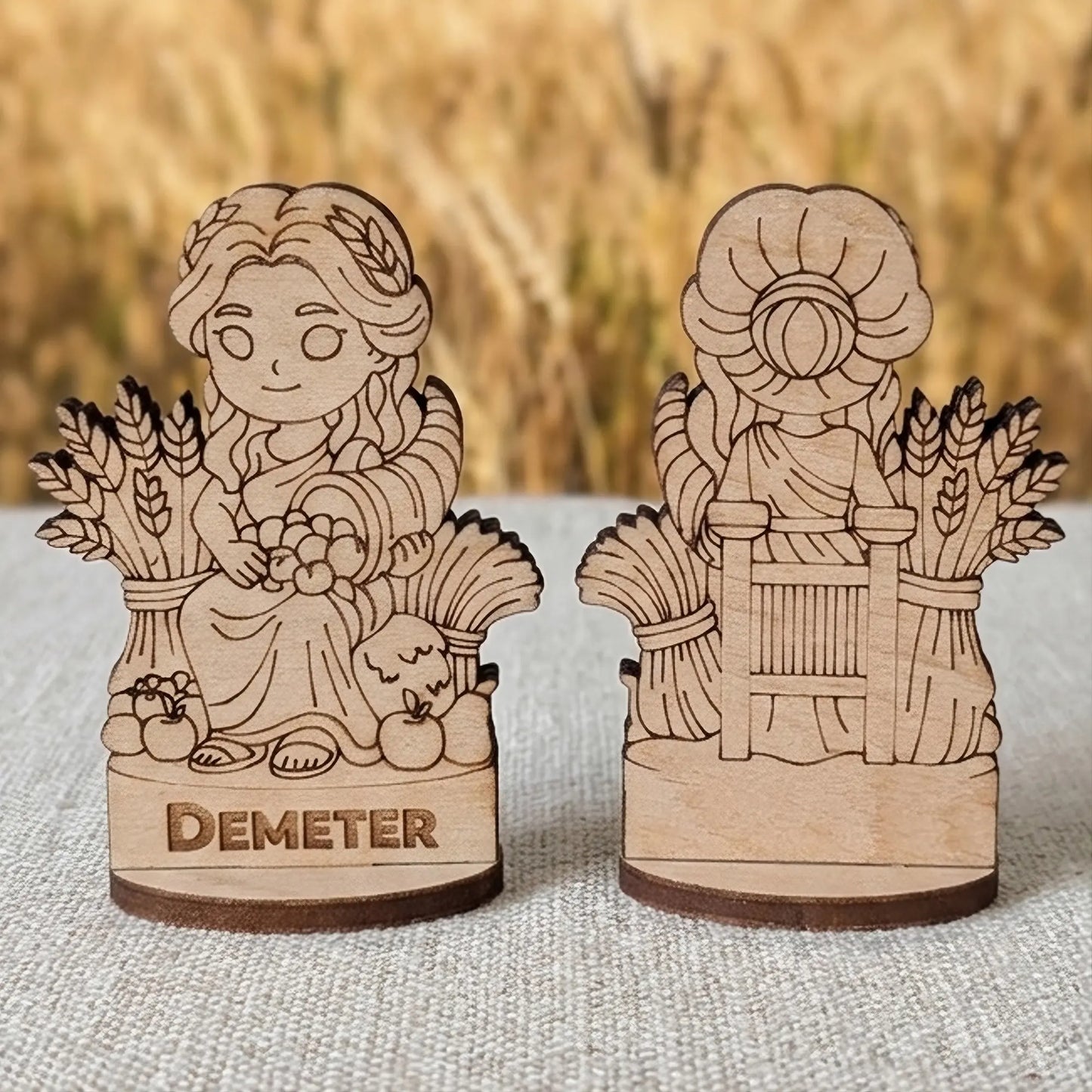 Front and back views of a laser cut plywood Demeter miniature with wheat and fruit against a blurry summer field background.
