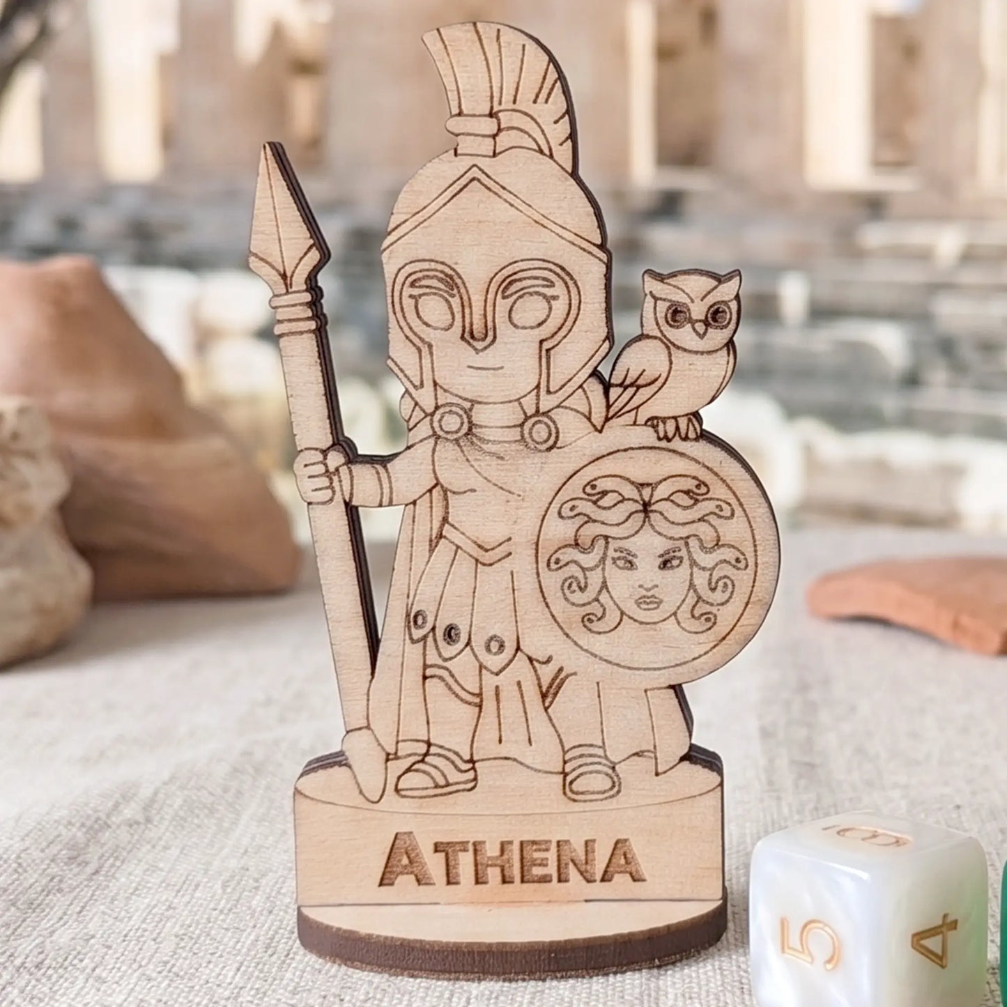Front view of laser cut plywood Athena miniature standing next to a white gaming die to show its small handcrafted scale.