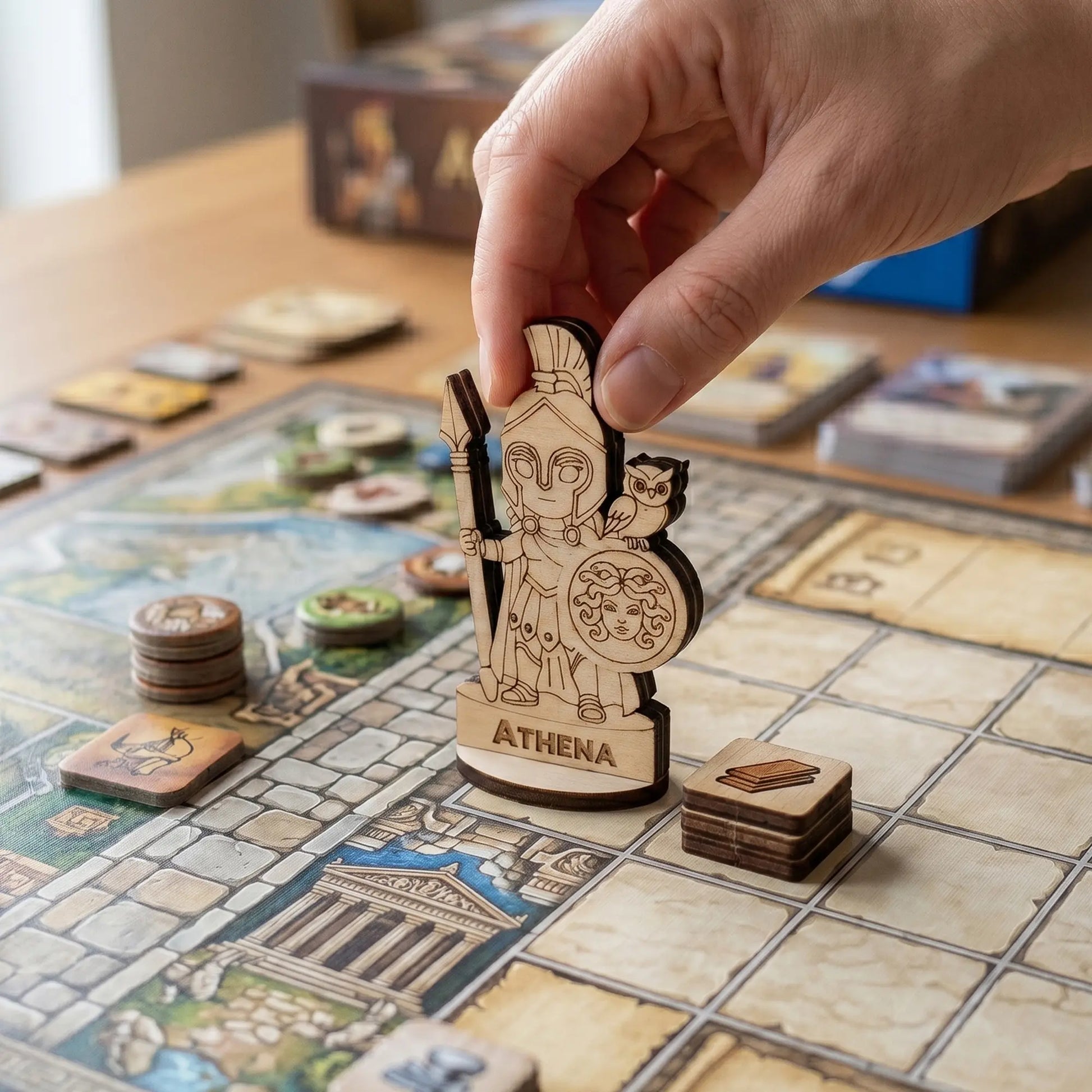An adult hand places a laser cut plywood Athena miniature on a grid board game map, surrounded by tokens and cards on a desk