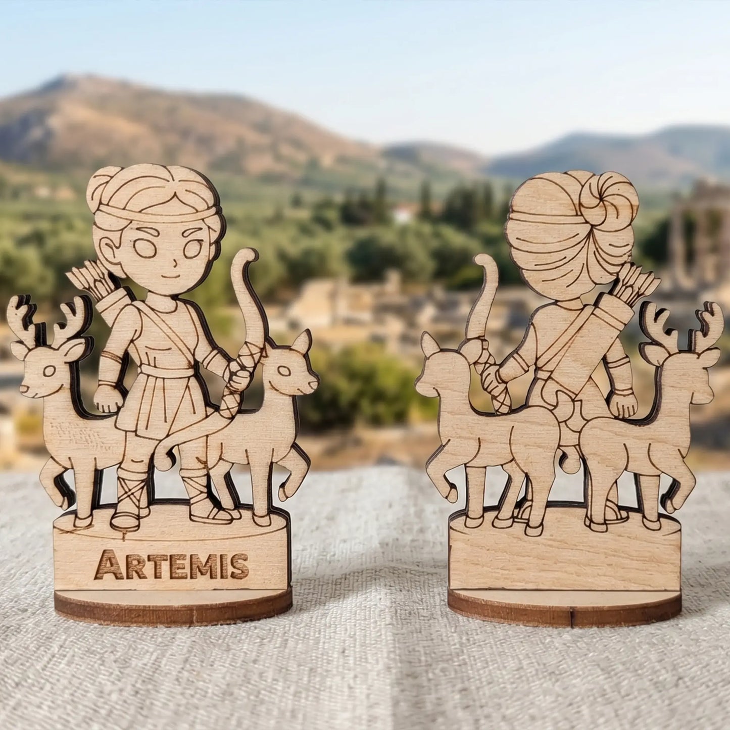 Front and back view of a laser cut plywood Artemis figurine with two deer and a bow, against blurred ancient Greek ruins