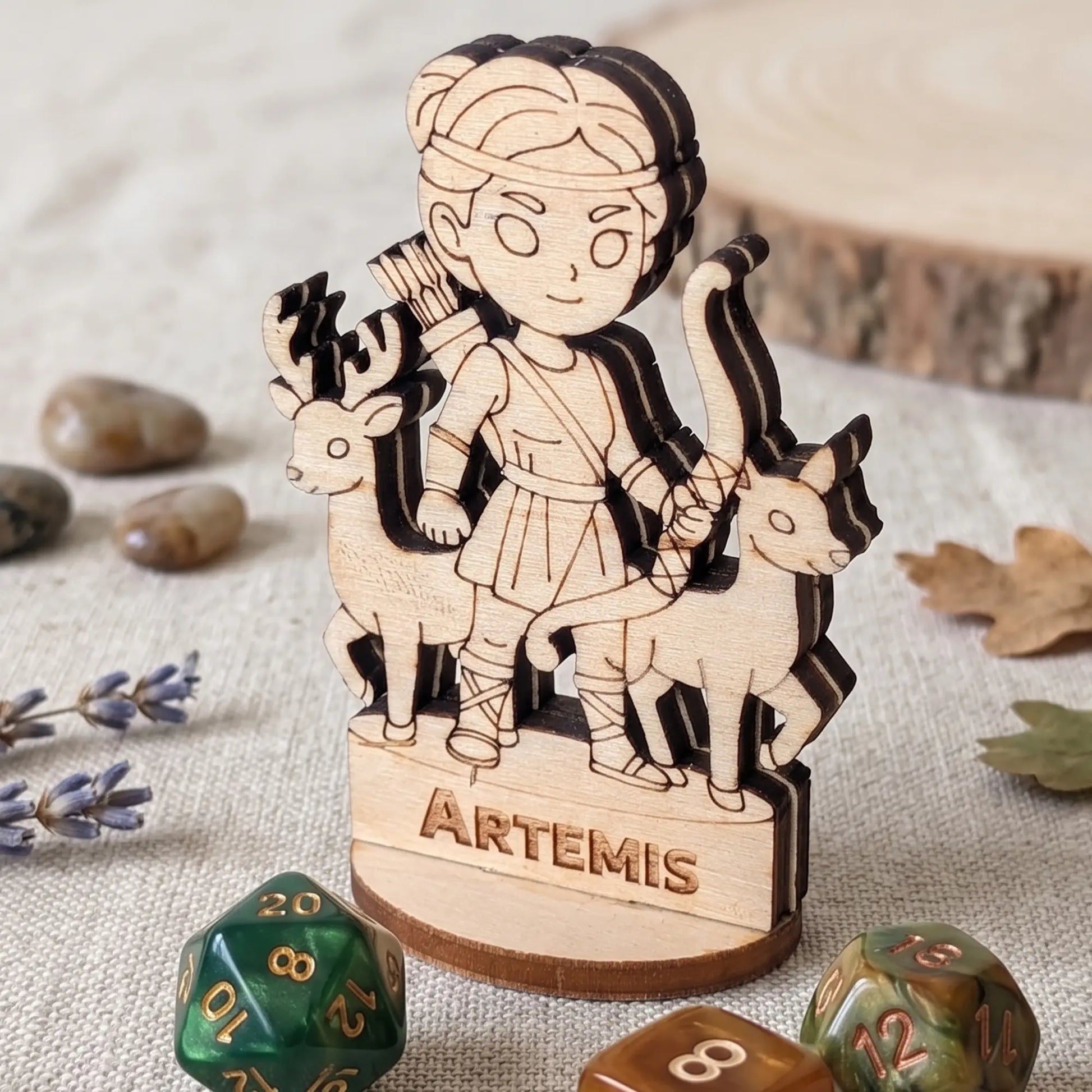 Laser cut plywood Artemis miniature on a table with green and brown polyhedral RPG dice, dried lavender, and smooth stones