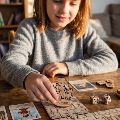 A child playing with a laser cut plywood Artemis miniature on a colorful tabletop RPG board game map with cards and dice