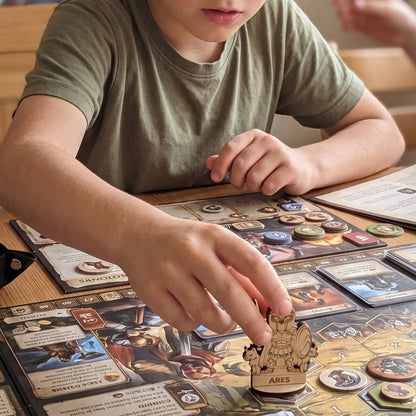 A child's hand moving a laser cut plywood Ares figurine across a colorful board game map with many tokens and game cards