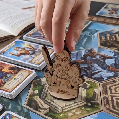 A hand carefully moves a laser cut plywood Ares miniature across a hexagonal board game tile map with various RPG cards