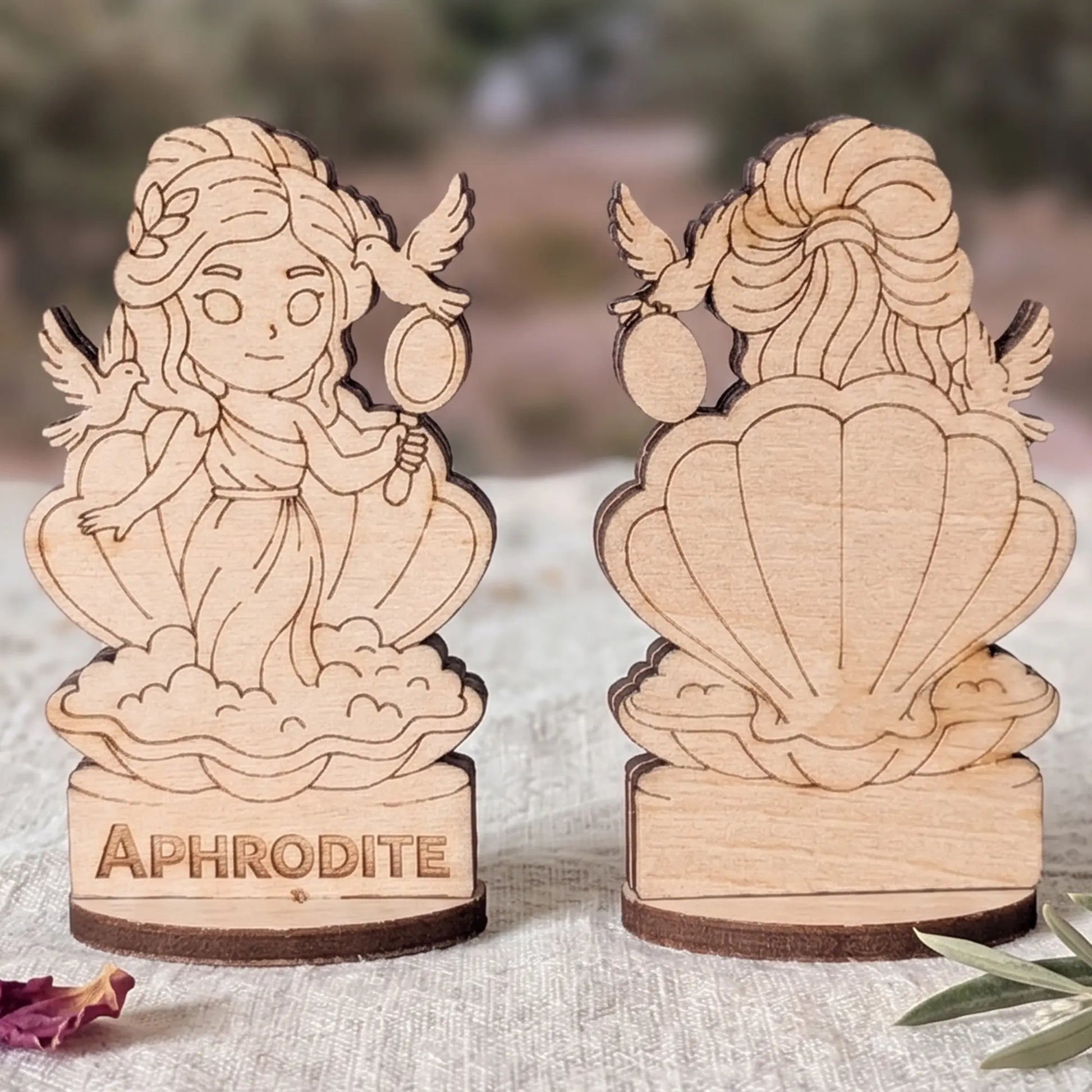 Front and back views of a laser cut plywood Aphrodite miniature in a shell with doves and mirror against blurred ruins