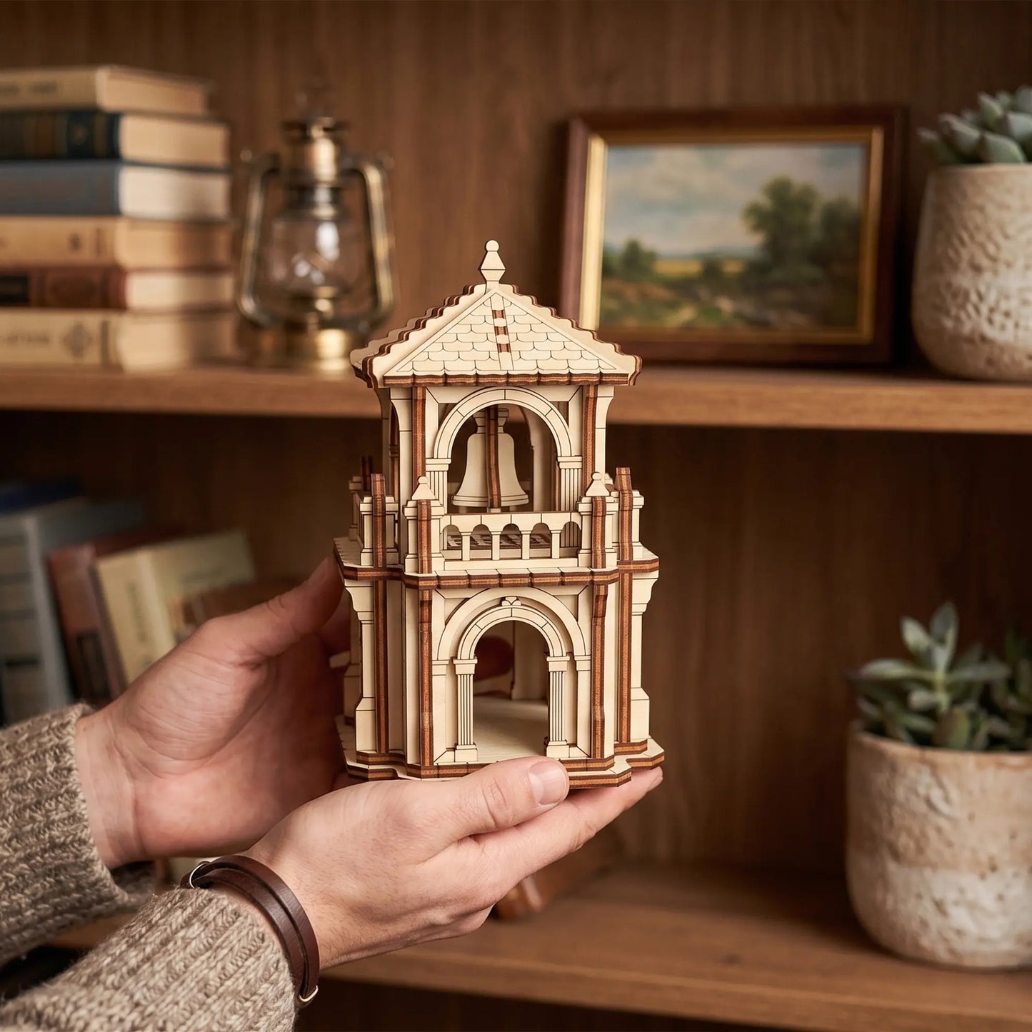 Hands holding a small fantasy bell tower puzzle model of laser cut plywood over wooden shelves filled with books.
