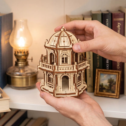 Hands holding a small laser cut plywood rotunda building on a bookshelf near books and a glowing vintage oil lamp.