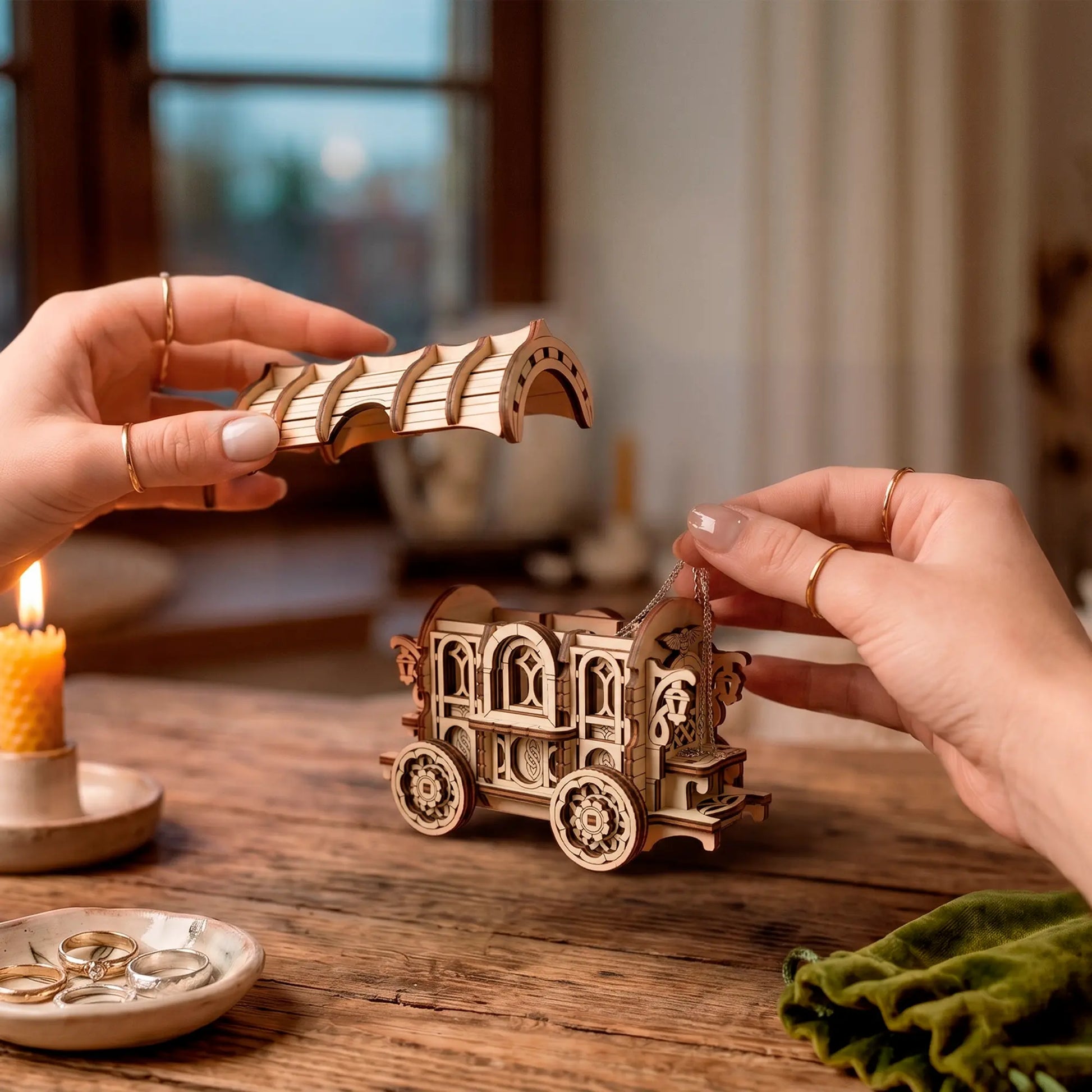 Hands with rings place the arched roof on a laser-cut wooden wagon puzzle box on a rustic table with a candle